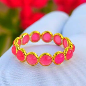 Pode incluir: Um anel dourado com uma série de pedras preciosas rosa vibrantes. As pedras preciosas são engastadas em biséis dourados individuais, criando uma faixa contínua de cor. O anel é exibido em uma superfície branca, com um fundo desfocado de flores vermelhas.