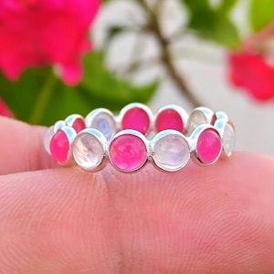 May include: A silver ring featuring alternating pink and clear gemstones. The ring is held on a finger. The background is blurred, with pink flowers and green leaves. The ring is a full eternity band.