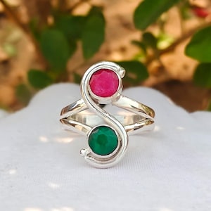 Può includere: Un anello in argento con un design distintivo. Presenta una gemma rosa brillante nella parte superiore e una gemma verde intenso nella parte inferiore, collegate da una fascia d'argento a spirale. L'anello è su uno sfondo bianco.