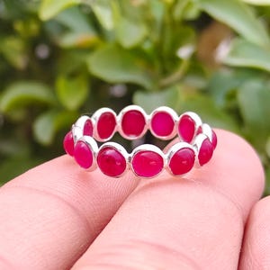 Puede incluir: Un anillo de plata con una serie de piedras preciosas rojas de forma ovalada. Las piedras están engastadas juntas, creando una banda continua de color. El anillo se sostiene sobre un fondo verde borroso.