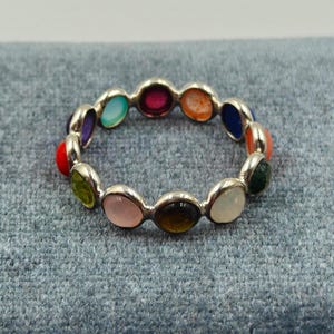 Puede incluir: Un anillo de plata con una serie de piedras preciosas ovaladas y coloridas. Las piedras preciosas están engarzadas en una fila alrededor del anillo, mostrando una variedad de colores, incluyendo rojo, rosa, verde y azul. El anillo se muestra sobre una superficie gris.