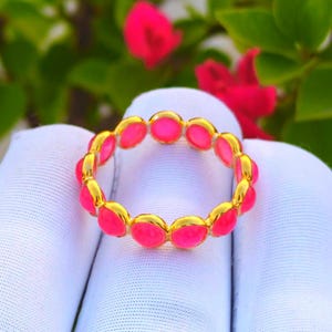 Könnte beinhalten: Ein goldfarbener Ring mit einer Reihe von rosa, oval geformten Steinen. Der Ring ist vor einem weißen Hintergrund mit grünem Laub und rosa Blüten platziert. Der Ring ist ein Schmuckstück.