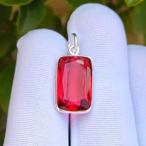 Könnte beinhalten: Ein rechteckiger, roter Edelsteinanhänger mit silberfarbenem Rahmen. Der Edelstein ist ein leuchtendes, transparentes Rot. Der Anhänger hat oben eine kleine Schlaufe für eine Kette. Der Hintergrund ist ein weißer Stoff.