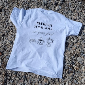 Könnte beinhalten: Weißes T-Shirt mit dem Text "REFRESH YOUR SOUL not your feed" und Illustrationen einer Zimtschnecke, einer Kaffeetasse und einer Kaffeekanne. Das T-Shirt liegt auf einem Bett aus kleinen Steinen.