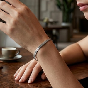 Op de afbeelding: Een zilverkleurige armband met visgraatketting gedragen om de pols. De armband heeft een plat, geweven metalen schakelontwerp met een gepolijste afwerking, wat zorgt voor een flexibele en strakke uitstraling die op de huid rust.