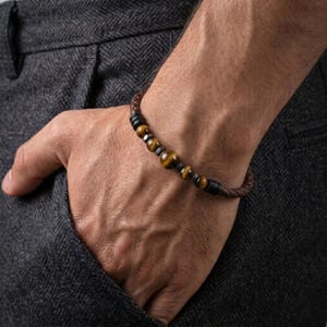 Op de afbeelding: Een armband met een gevlochten bruin leren koord, geaccentueerd met een reeks ronde tijgeroogkralen en kleine donkere metalen tussenstukjes. Het ontwerp bevat een grotere centrale tijgeroogkraal geflankeerd door kleinere kralen en zwarte accenten.