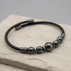 Può includere: Un braccialetto nero con perline grigio scuro, sferiche e sfaccettate. Le perline sono infilate su un cinturino nero flessibile. Il braccialetto è esposto su una superficie strutturata grigio chiaro.