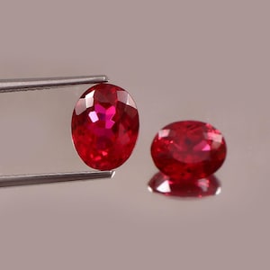 May include: Two oval-cut, vibrant red gemstones. One is held by silver tweezers, showcasing its facets. The other gem rests on a reflective surface, displaying its brilliant color and shape. The gems are a deep, rich red.