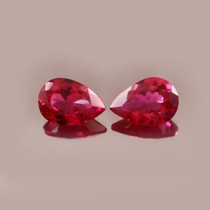Peut inclure: Deux pierres précieuses rouges en forme de poire, facettées, d'une grande clarté. Les gemmes sont présentées sur une surface réfléchissante, mettant en valeur leur brillance et leur couleur riche. Il s'agit probablement de rubis, un choix populaire pour les bijoux.