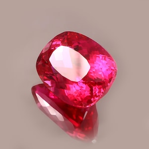 Puede incluir: Una gema de corte cojín con un color rojo intenso y saturado. La piedra presenta superficies facetadas complejas que crean reflejos geométricos nítidos y una apariencia brillante y centelleante en todo su cuerpo transparente.