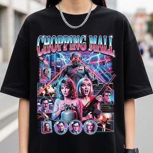 May include: Black t-shirt featuring a retro graphic design with the text "SHOPPING MALL" in a vibrant, multi-colored font. The design includes a robot, lightning bolts, and images of people, creating a vintage movie poster aesthetic.