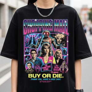 May include: Black t-shirt featuring a colorful graphic from the movie "Chopping Mall." The design includes the title in a gradient font, robot, and images of actors. Text includes "Buy or Die" and "Thank you, have a nice day!"