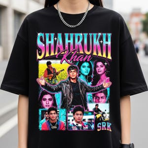 Puede incluir: Camiseta negra con un diseño gráfico retro de Shahrukh Khan. El diseño incluye el texto "Shahrukh Khan" y varias imágenes del actor. Se ve un collar de cadena plateada.