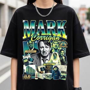 May include: Black t-shirt featuring a retro design with the name "MARK Corrigan" in large, stylized lettering. The design includes images of characters and text from a television show. The shirt has a crew neck and short sleeves.