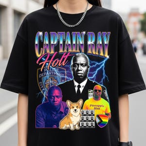 May include: Black t-shirt featuring a graphic design with the text "CAPTAIN RAY HOLT" in a retro style. The design includes portraits of a man, a corgi, and other elements. The shirt has a crew neck and short sleeves.