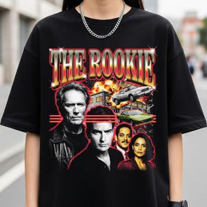 Può includere: T-shirt nero con un design retrò. Il testo "THE ROOKIE" è in rosso e oro. La grafica include immagini di attori e auto, con un'esplosione di fuoco sullo sfondo. Una t-shirt in stile vintage.