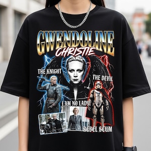 Puede incluir: Camiseta negra con un diseño gráfico que presenta el nombre "Gwendoline Christie" en letras doradas y azules. El diseño incluye imágenes de la actriz como caballero, diablo y en otros roles, con los textos "The Knight", "The Devil", "I Am No Lady" y "Rebel Scum".