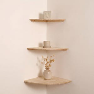 May include: Three wooden corner shelves mounted on a light-colored wall. The shelves hold a small ceramic face sculpture, a beige ceramic pot with a saucer, and a small vase with dried flowers. The shelves are unfinished wood.