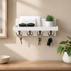 May include: A white wall-mounted shelf with four bronze-toned double hooks. The shelf holds a white envelope, a pair of black sunglasses, and a small potted succulent. Several metal keys and a black key fob hang from the hooks below the shelf.