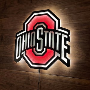 May include: Illuminated Ohio State logo sign. The sign features the words "OHIO STATE" in white, with a red "O" in the centre. The sign is outlined in black and is mounted on a wooden wall.
