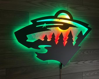 Pode incluir: Uma peça de arte de parede em metal preto com iluminação LED verde e vermelha. O design inclui uma estrela, árvores e um elemento circular, com um fundo branco. A peça é montada numa parede de madeira.