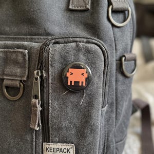 Op de afbeelding: Een ronde button met een gestileerd oranje pixelkarakter op een zwarte achtergrond, bevestigd aan het voorvak van een donkergrijze canvas rugzak met een rits en een label met de tekst KEEPACK.