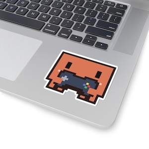 May include: A square-shaped pixel art sticker featuring a light orange character with two black eyes and pink cheeks, holding a dark grey video game controller with small red, yellow, green, and blue buttons, displayed on a silver laptop surface.