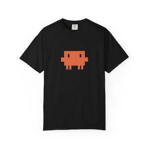 May include: Black t-shirt featuring a pixelated orange creature design. The graphic is centered on the front of the shirt. The t-shirt has short sleeves and a crew neck. The fabric appears to be a soft cotton blend.