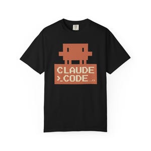 May include: Black t-shirt featuring a pixelated design with a brown figure above the text "CLAUDE >_CODE...". The design has a retro video game aesthetic. The shirt is made of a soft material.