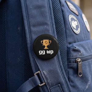 May include: A circular pinback button with a black background featuring a pixelated gold trophy icon and the white pixelated text "gg wp" centered below the trophy. The button is attached to the dark blue fabric strap of a backpack.