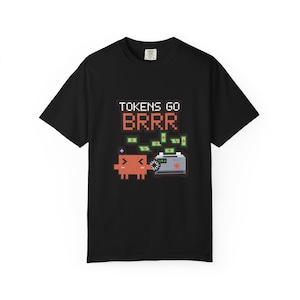 May include: A black short-sleeve t-shirt featuring a pixel art graphic of a small orange character next to a toaster emitting green money. The text above the graphic reads TOKENS GO BRRR in a pixelated font. The design is centered on the chest.