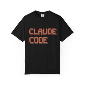 May include: Black t-shirt with the words "CLAUDE CODE" in a pixelated, brick-like font. The text is in a reddish-brown color and is centered on the front of the shirt. The shirt has short sleeves and a crew neck.