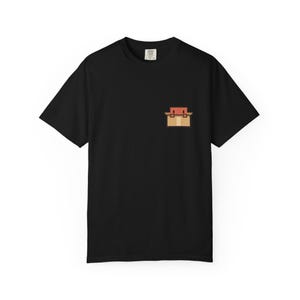 May include: A black short-sleeved t-shirt featuring a small pixel art graphic on the left chest. The graphic depicts a tan cardboard box with a red square-shaped character peeking out from the top. The shirt has a standard crew neckline.