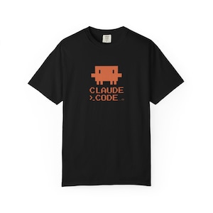Op de afbeelding: Zwart T-shirt met een oranje pixelachtig figuur en de tekst "CLAUDE >.CODE..". Het shirt met korte mouwen heeft een eenvoudig ontwerp met de afbeelding in het midden.