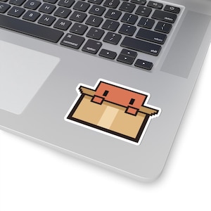 May include: A die-cut sticker featuring a pixel art design of a tan cardboard box with a coral-colored lid. The box has a simple, blocky aesthetic with dark brown outlines and a light tan vertical stripe down the center of the front panel.
