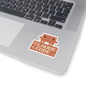 May include: A sticker featuring a pixelated brown robot holding a sign that reads "CLAUDE >.CODE...<". The sticker is rectangular with a white border and is placed on a laptop.