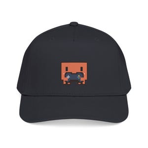 Puede incluir: Una gorra de béisbol gris oscuro con un gráfico de pixel art centrado que muestra un personaje cuadrado de color naranja sosteniendo un pequeño mando de videojuego azul oscuro. El diseño está compuesto por píxeles cuadrados.