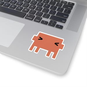 Puede incluir: Una pegatina troquelada que muestra un personaje pixelado y cuadrado de color naranja apagado. El personaje tiene un cuerpo rectangular con cuatro patas cortas y dos ojos negros pequeños en forma de V lateral que parecen estar cerrados.