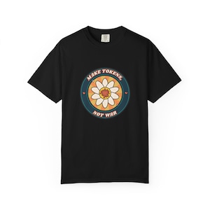 May include: A black short-sleeved t-shirt featuring a circular graphic on the chest. The design includes a white daisy flower over an orange sunburst background, surrounded by the text "MAKE TOKENS, NOT WAR" in a dark blue circular border.