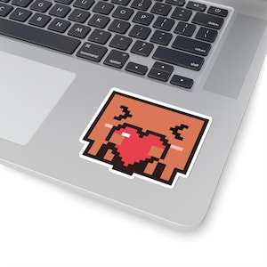 Könnte beinhalten: Ein quadratischer Pixel-Art-Aufkleber mit einer hellbraunen Figur mit fröhlichem Ausdruck, geschlossenen Augen und einem markanten roten Herz in der Mitte. Das Design verwendet eine blockartige Retro-Ästhetik mit weißem Rand auf einem Laptop.