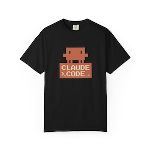 May include: Black t-shirt with a pixelated graphic of a figure above the text "CLAUDE >.CODE...". The design is in shades of orange and tan. The shirt is a short-sleeved crew neck.