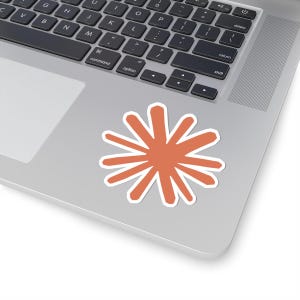 May include: A sticker with an orange burst design and a white border, positioned on a silver laptop. This sticker could be used to decorate a device. The design is simple and modern. The sticker's shape resembles a starburst.