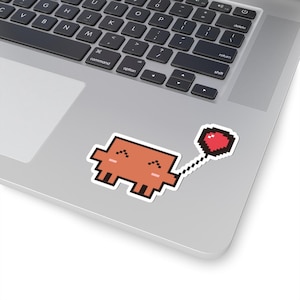 May include: A pixel art sticker featuring a rectangular orange creature with a smiling face, holding a red heart-shaped balloon on a string. The sticker is applied to the surface of a silver laptop with a black keyboard.
