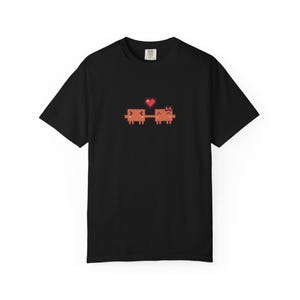 May include: A black short-sleeve t-shirt featuring a pixel art graphic of two orange square-shaped characters holding hands. Above the characters is a small red heart. The shirt has a crew neckline and a standard fit.