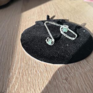 Könnte beinhalten: Ein silberfarbenes, drahtgewickeltes Schmuckset mit einem Armband und einem Anhänger, jeweils mit einem hellgrünen Edelstein. Das Set liegt auf einem schwarzen Samtbeutel, vor einem hellen Holzmaserungshintergrund.