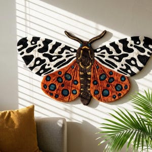 May include: A large moth-shaped rug, ideal for wall decor. The wings display a striking pattern of black and white, with orange lower wings adorned with blue spots. The rug is mounted on a white wall, adding a unique touch to the room's decor.