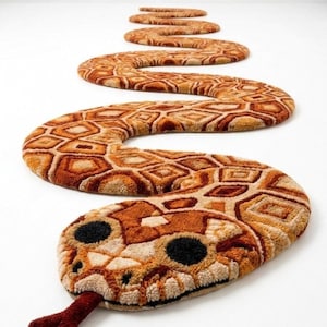 May include: A long, snake-shaped rug featuring a textured, tufted surface with a repeating geometric pattern in shades of brown, tan, and cream. The rug has a rounded head with two large black circular eyes and a protruding dark red forked tongue.