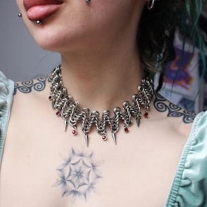 May include: A silver chainmail choker necklace with red accents and dangling spikes. The necklace is made of interlocking metal rings and features small red beads. The necklace is worn around the neck.