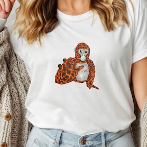May include: White t-shirt featuring a cartoon orangutan design. The orangutan has a textured orange and black pattern on its body and a light gray chest. The design is centered on the front of the shirt.