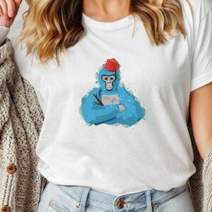 May include: White t-shirt featuring a blue watercolor illustration of a monkey wearing a red fez and holding a stick. The monkey has its arms crossed and the word "electronic" is printed on a small sign.